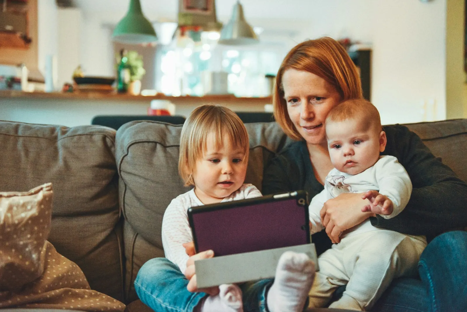 A parent and two small children looking at a tablet together on a sofa